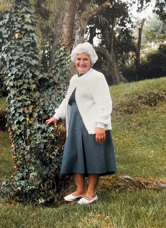 Elisa, white-haired, in a white cardigan, her hand resting on an ivy-covered tree